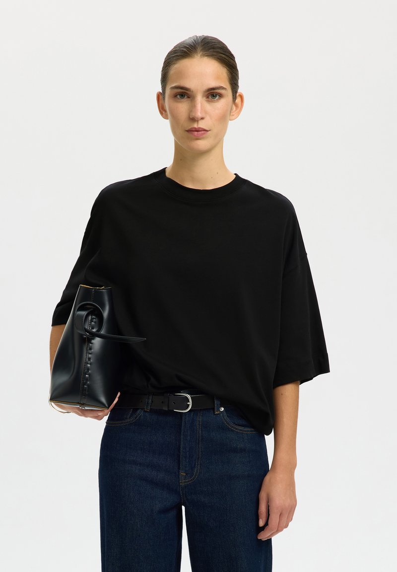 Woman with slicked-back hair wears a black oversized t-shirt and dark jeans, holding a black leather handbag with a top handle.