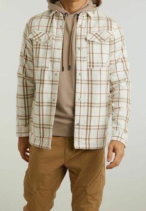 Homme portant un sweat à capuche beige sous une chemise à carreaux blanche et marron, associée à un pantalon cargo marron clair, debout les bras détendus.