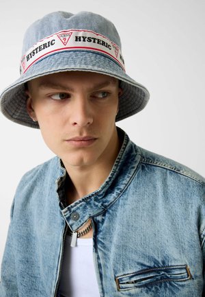 Młody mężczyzna noszący jasny jeansowy kapelusz typu bucket z czerwonymi logo "HYSTERIC" i "GUESS", jasną jeansową kurtkę oraz srebrny łańcuszek na szyi.