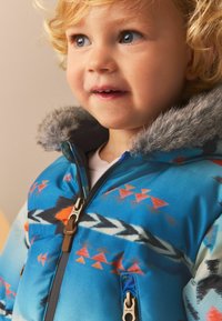 Chaqueta azul para niños con patrones tribales en rojo y negro. Cuenta con una capucha forrada de piel y dos bolsillos con cremallera. Textura suave y brillante.