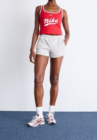 Červený bezrukávový top s bílým logem Nike, spárovaný se světle šedými šortkami a bílými ponožkami. Obuv má červené a šedé akcenty.
