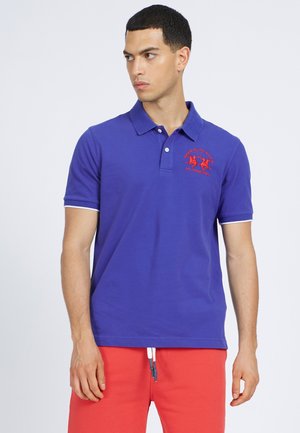 Polo shirt viola con colletto, maniche corte e un logo ricamato rosso sul petto. Indossato con pantaloni corti rossi con lacci bianchi.