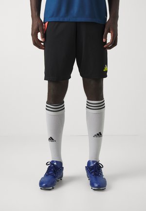 Zwarte sportshorts met een neongele logo, gecombineerd met witte kniekousen met zwarte strepen en blauwe voetbalblessers.
