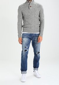 Pull gris en maille avec motif torsadé texturé, col à deux boutons ; porté avec un jean bleu déchiré et des baskets blanches.