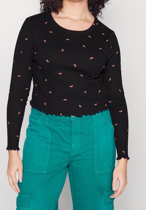 Personne portant un haut noir à manches longues avec de petites impressions arc-en-ciel et un pantalon taille haute bleu sarcelle avec poches avant et latérales.