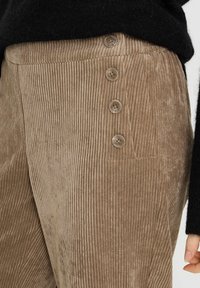 Pantalones de pana marrones con costillas verticales, bolsillo lateral y cuatro grandes botones en la cintura. Textura suave con un ligero brillo.