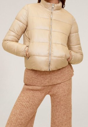 Veste matelassée beige avec fermeture éclair à l'avant, portée par-dessus un pull en tricot côtelé camel et un pantalon large assorti.