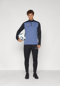 Nike sportswear sæt bestående af en blå langærmet top med sorte ærmer og lynlås, kombineret med sorte tætsiddende bukser og sorte sportssko.