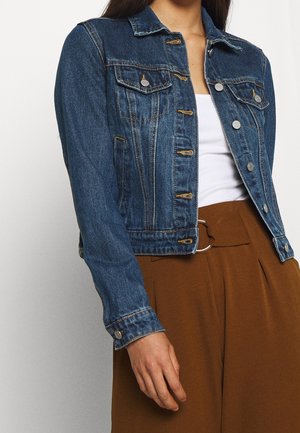 Femme portant une veste en jean bleue sur un haut blanc, associée à un pantalon marron taille haute avec une boucle de ceinture argentée en forme d'anneau.