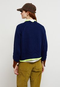 Femme aux cheveux roux portant un pull en maille bleu marine avec une bordure néon, des manches marron et orange, un pantalon olive et une casquette à carreaux marron, vue de profil.