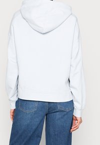 Sudadera corta de color azul claro con capucha, dobladillo acanalado y corte holgado. El material parece suave; presenta hombros caídos y no tiene gráficos visibles.