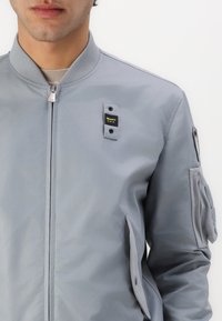 Šviesiai pilkas bomber stiliaus švarkas su ripsu apykakle, priekiniais užtrauktukais ir šoninėmis kišenėmis. Yra logotipo aplikacija kairėje krūtinės pusėje ir užtrauktuko kišenė ant rankovės.