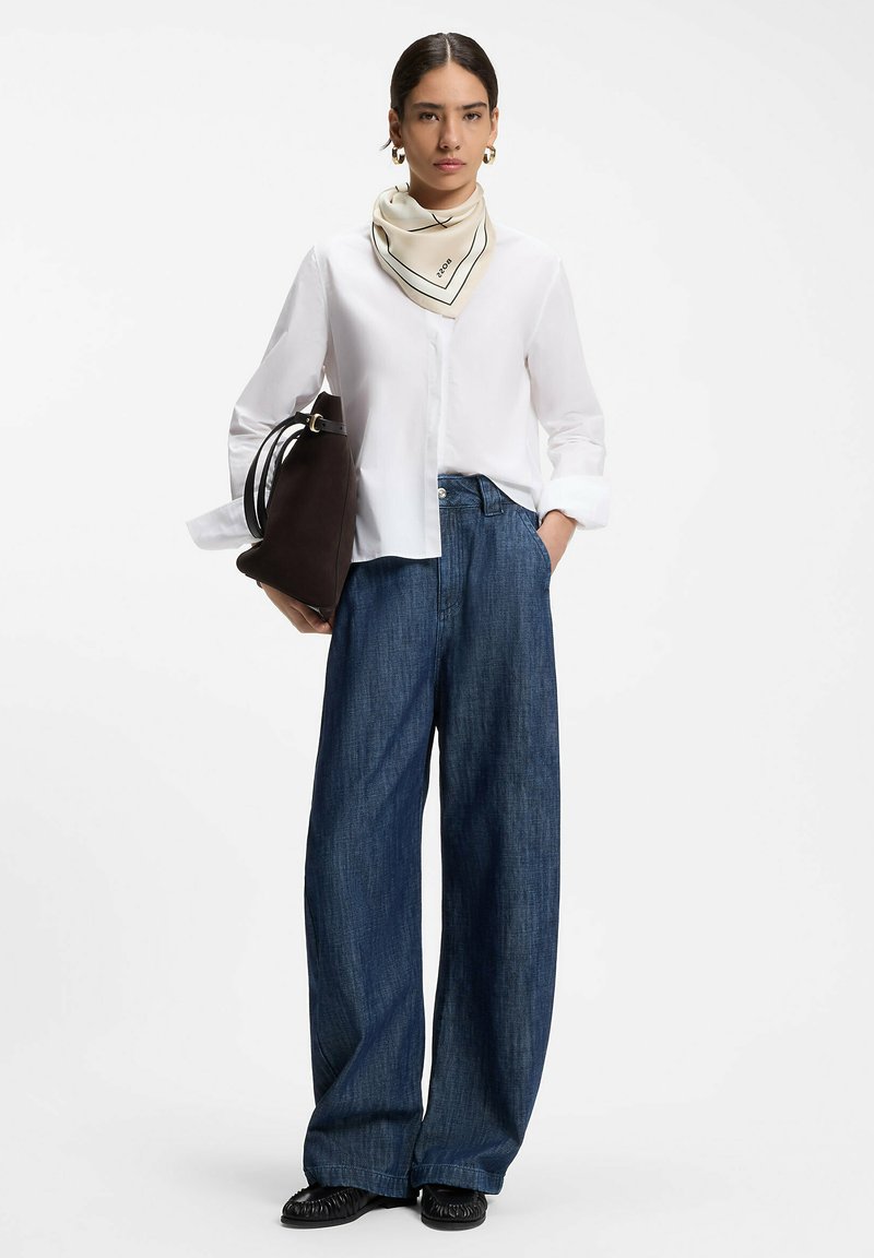 Femme portant un chemisier blanc, un jean bleu à jambes larges, des chaussures noires, une écharpe crème, des boucles d'oreilles créoles dorées, tenant un grand sac à main noir.