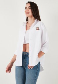 White button-up shirt made of cotton, featuring a small brown bear embroidery on the left side, paired with blue denim jeans.
