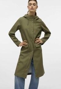 Olivno zelena vodoodporna jakna s kapuco, z gumbi na zapiranje, dvema sprednjima žepoma in dolgimi rokavi, kombinirana s svetlim denim jeansom.