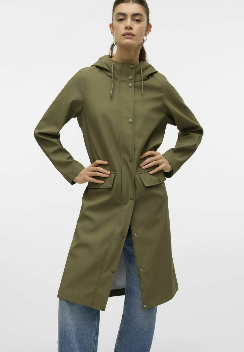 Olivno zelena vodoodporna jakna s kapuco, z gumbi na zapiranje, dvema sprednjima žepoma in dolgimi rokavi, kombinirana s svetlim denim jeansom.