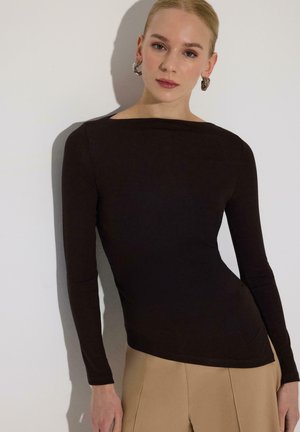 Femeie cu piele deschisă și păr blond poartă un top negru mulat cu mâneci lungi, pantaloni bej și cercei argintii în formă de cerc, stând lângă un perete alb.