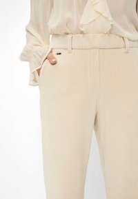 Pantalons en velours côtelé beige avec une coupe droite, tissu texturé, poches latérales et un détail de logo discret sur la ceinture.