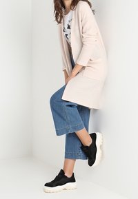 Hellrosa langer Cardigan in entspannter Passform, getragen über einem grafischen T-Shirt. Weite Denim-Culottes und schwarze Sneakers mit weißen Sohlen vervollständigen den Look.