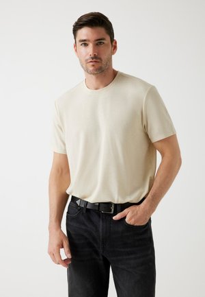 Beige, rövid ujjú póló kerek nyakkivágással, fekete farmernadrággal és fekete övvel viselve. Simaság jellemzi, minimális dizájn, minták vagy grafikák nélkül.