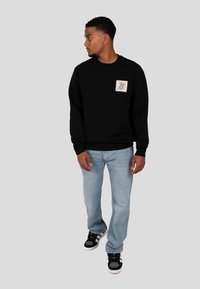 Pullover nero con collo a giro e grafica sulla tasca frontale, abbinato a jeans blu chiaro e sneakers nere. Il tessuto sembra morbido e rilassato.