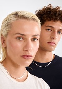 To personer i nærbillede, den ene med platinblond hår og en beige skjorte, den anden med krøllet brunt hår i en marineblå top, begge iført sølvhalskæder.