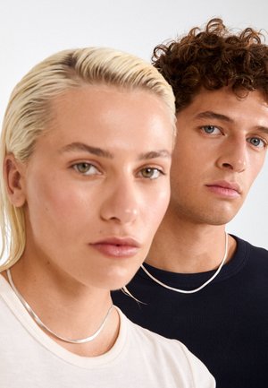 Two individuals in close-up, one with platinum blonde hair and a beige shirt, the other with curly brown hair in a navy top, both wearing silver necklaces.