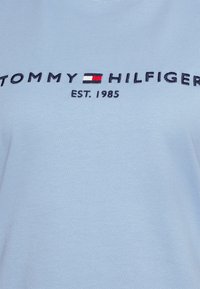 Tessuto azzurro chiaro con testo ricamato "TOMMY HILFIGER" e il logo della bandiera rossa, bianca e blu del marchio, con "EST. 1985" sotto.