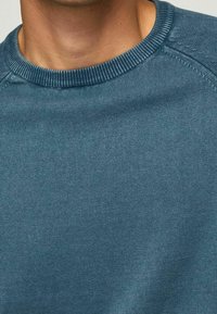 Detailní záběr osoby v modrém pleteném svetru s kulatým výstřihem, zaměřující se na výstřih a horní část hrudníku.