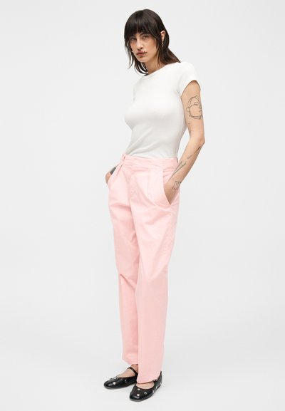 Femme tatouée porte un haut blanc ajusté à manches courtes, un pantalon taille haute rose clair et des ballerines noires à clous argentés, mains dans les poches.