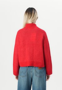 Persoon met lang blond haar, gekleed in een rode coltrui en losse blauwe spijkerbroeken, gezien vanuit de rug tegen een witte achtergrond.