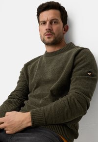 Mørkegrøn strikket sweater med ribbet kant ved ærmer og bund. Har en lille logoapplikation på ærmet. Bæres over en skjorte, afslappet pasform.