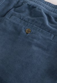 Primo piano della tasca di pantaloni in velluto a coste blu con un bottone marrone e una cintura elastica visibile nella parte superiore.