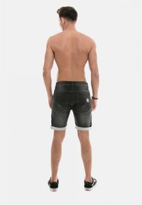 Shorts en denim noir avec des ourlets roulés, dotés de poches arrière et d'un patch logo circulaire, vus de l'arrière. Le matériau est texturé et légèrement décoloré.