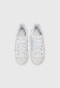 Ténis brancos com riscas acentuadas em azul claro, biqueira em borracha texturizada e atacadores planos brancos. Parte superior sintética lisa com colarinho acolchoado.