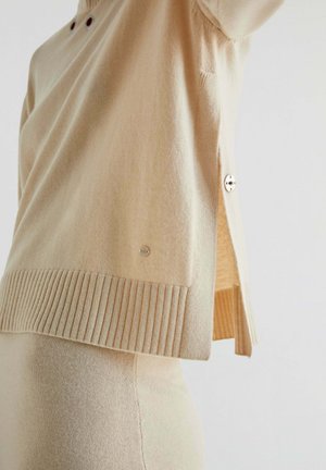 Beige strikket sweater med ribbet kant og sideslidser. Har knapdetaljer og en glat tekstur. Blød, afslappet stil der er velegnet til lag-på-lag.