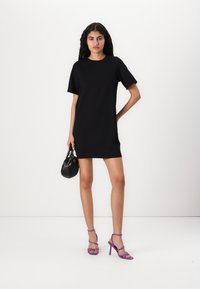 Rochie neagră cu mâneci scurte și croi lejer. Modelul ține o geantă mică neagră și poartă sandale cu toc înalt, cu bretele, de culoare violet. Textură netedă.