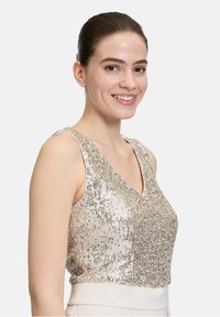 Skrząca się crop top z cekinami w srebrnych i złotych odcieniach, z dekoltem w kształcie litery V i bez rękawów, tworzący teksturowaną, refleksyjną powierzchnię.