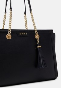 Black leather handbag with gold chain straps, DKNY logo, and a black tassel detail. Smooth texture with structured design and accents.
