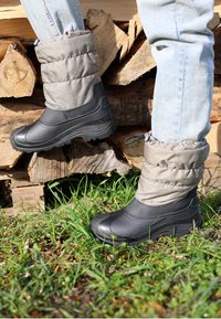 Schwarze wasserdichte Stiefel mit grauen, gesteppten Bündchen, einer strukturierten Gummisohle und glattem Obermaterial, auf Gras und Holz platziert.