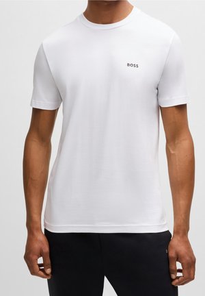 Mand iført en enkel hvid kortærmet t-shirt med "BOSS" logo på venstre bryst og sorte bukser mod en neutral baggrund.