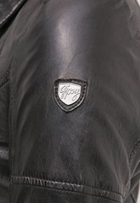 Giacca di pelle nera con finitura testurizzata, cuciture visibili e un logo a forma di scudo argentato sulla spalla sinistra.