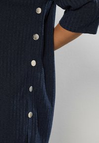 Giarmento lavorato a maglia blu navy con un motivo testurizzato e bottoni a pressione in tonalità argento lungo il lato. Maniche corte e design morbido.