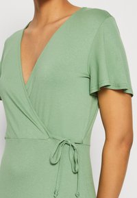 Vihreä kietaisumekko lyhyillä lentosarvilla, syvä V-pääntie, sitomisdetalji vyötäröllä, valmistettu sileästä, venyvästä kankaasta.