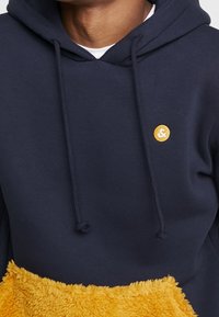 Marinblå hoodie med en gul, mjuk framficka och en liten rund gul logotyp på bröstet, bärs över en vit skjorta.