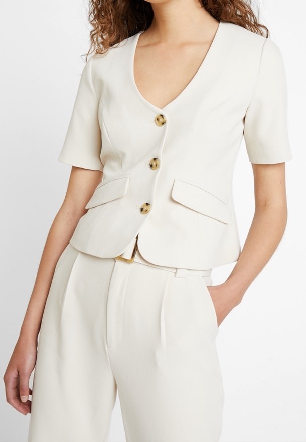 Femme portant un blazer crème à manches courtes avec trois boutons et un pantalon taille haute assorti, main dans la poche, sur fond blanc.