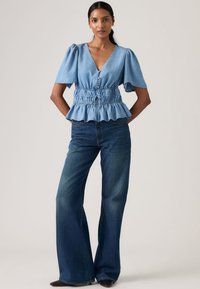 Ανοιχτό μπλε τοπ πεπλούμ με κοντά φουσκωτά μανίκια και V-λαιμόκοψη, σε συνδυασμό με σκούρο μπλε παντελόνι φαρδιάς γραμμής με ξεθωριασμένη υφή.