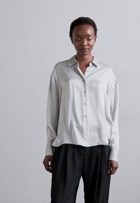 Ανοιχτό γκρι σατέν πουκάμισο με κουμπιά, γιακά, μακριά μανίκια και χαλαρή εφαρμογή, σε συνδυασμό με μαύρους παντελόνες. Λείο υλικό και μίνιμαλ σχέδιο.