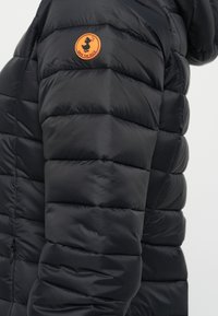 Zwarte pufferjack met een doorgestikte textuur, voorzien van een oranje, rond logo op de linker schouder, ontworpen voor warmte en isolatie.