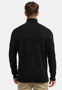 Svart stickad tröja med hög krage, med texturerade horisontella ränder och ribbade ärmslut. Materialet är mjukt med medelhög tjocklek.